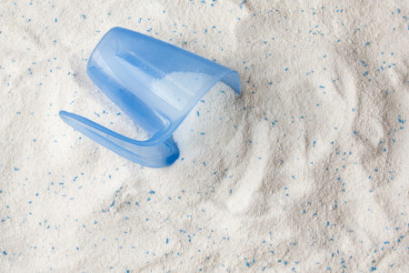 Waschmittel bzw. Waschpulver. Weißes Pulver mit einigen blauen Partikeln. Darauf liegt ein Dosierbehälter