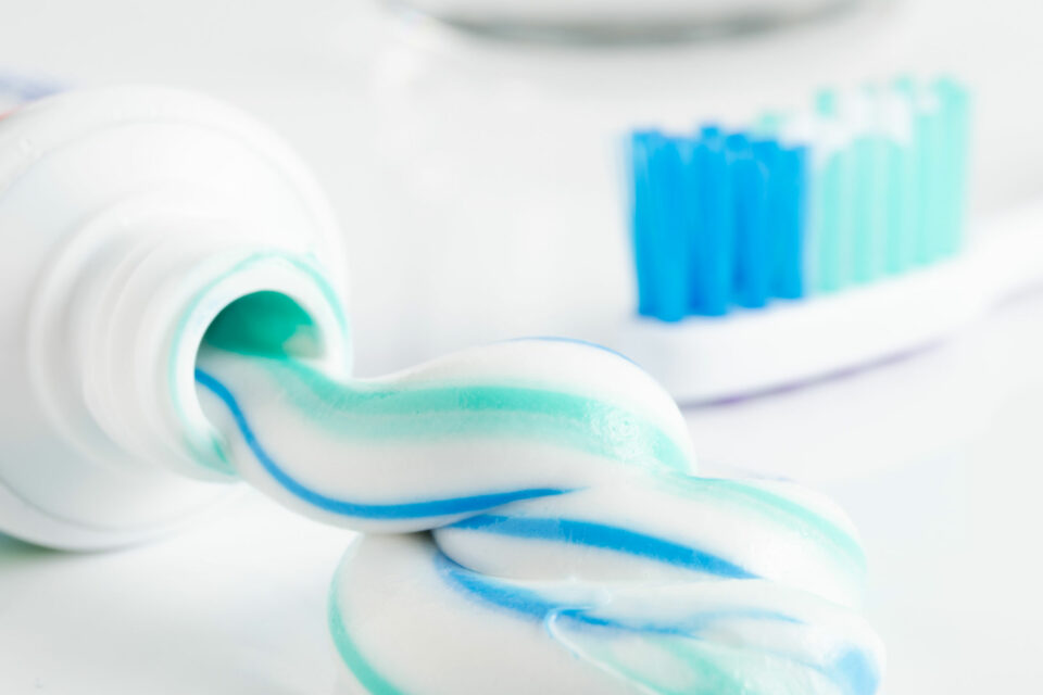 Eine offene Tube Zahnpasta und eine Zahnbürste liegen auf einem Waschtisch. Aus der Tube quillt blau-weiße Zahnpasta